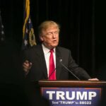 Man speaking at podium with "TRUMP" sign.