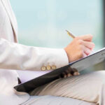 Person in beige suit writing on a clipboard.