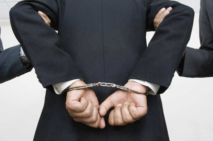 Person handcuffed in suit, escorted by two others.