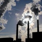 Silhouetted smokestacks emitting smoke against a cloudy sky with sunlight