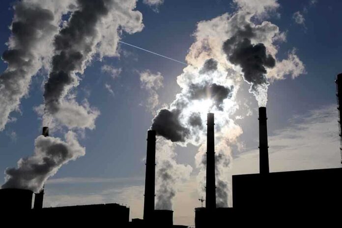 Silhouetted smokestacks emitting smoke against a cloudy sky with sunlight