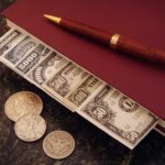 A stack of cash and coins on a book with a pen