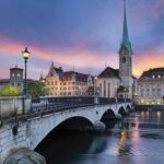 Catholic Event Shock: Clergy FURIOUS! Scenic view of Zurich with a bridge, historic buildings, and a sunset sky