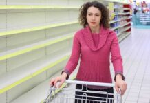 Grocery Prices Set To HIKE After Bill Passed A woman with curly hair stands in a grocery store aisle with empty shelves, pushing a shopping cart.
