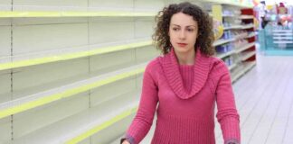 A woman with curly hair stands in a grocery store aisle with empty shelves, pushing a shopping cart.
