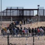 Crowd of people lining up near a border fence with barbed wire