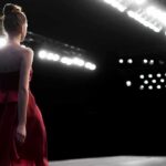 Model in a red dress walking on a runway under bright stage lights