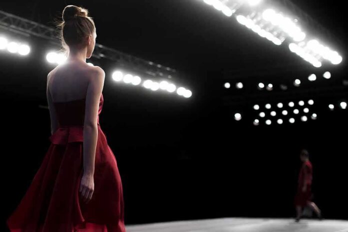 shutterstock_370693958 Model in a red dress walking on a runway under bright stage lights
