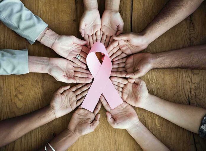 shutterstock_589644902 Hands forming a circle around a pink ribbon on a wooden table