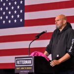 Man in hoodie speaking at a podium before a large U.S. flag