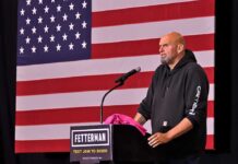 Man in hoodie speaking at a podium before a large U.S. flag
